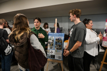 Kilka osób stoi przy stoisku w jasnym pomieszczeniu. Na pierwszym planie widać tablicę na sztaludze z niebieskim plakatem zawierającym zdjęcia i napisy o kole naukowym. Jedna osoba w zielonej bluzie rozmawia z osobą w ciemnoszarej koszulce, obok stoi osoba w białej koszuli. 