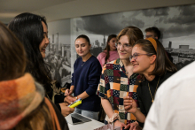 Kilka osób stoi przy stole w jasnym pomieszczeniu. Na pierwszym planie widać osobę trzymającą żółty marker. Na stole leży laptop, ulotki i małe przedmioty. Jedna osoba ma na sobie koszulę w kolorowe geometryczne wzory, obok stoi osoba w granatowym swetrze. W tle widoczna jest czarno-biała fotografia miasta na ścianie.