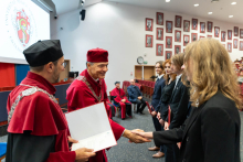 Dwóch mężczyzn w czerwonych togach i biretach wręcza dyplomy trzem młodym osobom stojącym w rzędzie. Jeden z nich uśmiecha się i podaje dyplom, drugi trzyma otwartą teczkę. W tle widać siedzące osoby. Po lewej stronie ekranu wyświetla się logo wydziału.