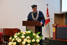 Dziekan wydziału. Mężczyzna w granatowym stroju akademickim i czarnym birecie stoi za mównicą. Przemawia do zgromadzonych. W tle widać flagę z orłem i napis „VERITATIS SPLENDOR”. Przed mównicą stoi bukiet białych róż z zielonymi liśćmi.