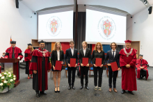 Na scenie stoi sześć osób. Cztery osoby, immatrykulowani studenci, są w środku i trzymają czerwone teczki. Po bokach stoją mężczyźni w czerwonych togach i biretach. Są to dziekan i prorektor.