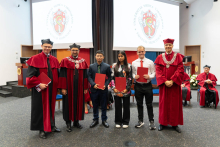 Na scenie stoi sześć osób. W środku są studenci, ubrani formalnie. Trzymają czerwone teczki. Po bokach stoją mężczyźni w czerwonych togach z insygniami rektorskimi i dziekańskimi. 