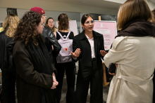 Kilka osób stoi w jasnym pomieszczeniu przy stoisku informacyjnym. Na pierwszym planie widać osobę w jasnym płaszczu trzymającą telefon, naprzeciwko stoi osoba w czarnym garniturze z rozłożonymi rękami. Obok stoją osoby w ciemnych ubraniach, jedna ma plecak w kolorze jasnym z czerwonym logo. W tle znajdują się dwa plakaty na sztalugach z tekstem.