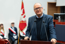 Mężczyzna w średnim wieku, prof. Leszek Czupryniak, przemawia przy mównicy. Nosi granatową marynarkę, niebieską koszulę i okulary. Przemawia do mikrofonu.