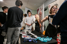 Kilka osób stoi przy stoisku w jasnym pomieszczeniu. Na stole leżą ulotki, stetoskop w niebieskim kolorze, zielony fartuch i małe przedmioty medyczne. Jedna osoba w czarnej sukience gestykuluje, a obok stoją osoby w jasnych i ciemnych ubraniach. 