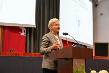 Senator Małecka-Libera. Kobieta w średnim wieku z krótkimi, jasnymi włosami stoi przy mównicy i przemawia. Nosi szary żakiet i białą bluzkę. Gestykuluje rękami. W tle widać duży ekran z logo uczelni medycznej oraz czerwony baner z symbolem uczelni. 