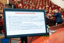 a pierwszym planie widać monitor z tekstem zatytułowanym „Dekalog pierwszoroczniaka”. Po prawej stronie dwie osoby stoją przy mównicy. Tekst zawiera dziesięć rad dla studentów pierwszego roku medycyny.