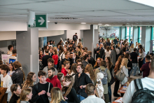 Kilkadziesiąt osób stoi w korytarzu przy stoiskach edukacyjnych. Ludzie rozmawiają i oglądają materiały na stołach. Widać manekin anatomiczny z białym szkieletem. Na ścianach wiszą plakaty, a w tle są duże okna z widokiem na zieleń. 