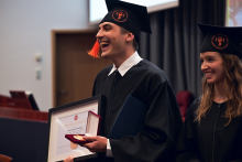 Osoba ubrana w czarną togę akademicką i biret z pomarańczowym chwostem stoi, trzymając w dłoniach ramkę z dyplomem oraz pudełko z medalem. 