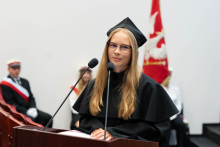 Młoda kobieta, przewodnicząca samorządu WL, w czarnej todze i birecie stoi za mównicą i przemawia do mikrofonu. Ma długie blond włosy i okulary. W tle widać trzy osoby w galowych mundurach oraz biało-czerwoną flagę z orłem. 