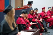 Po lewej stronie młoda kobieta, przewodnicząca samorządu WL, przemawia do mikrofonu. Obok siedzi siedem osób w czerwonych togach i biretach. 