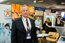 A man, Prof. Marcin Sobczak, wearing a navy blue suit, stands in front of an exhibition stand with the CEPT logo and a photo of a person in a laboratory. In the background, several people are looking through materials on tables. 