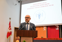 Prezes Nowak. Mężczyzna w ciemnym garniturze i okularach stoi za mównicą i przemawia. Za nim wisi ekran z napisem „Immatrykulacja studentów kierunków Wydziału Nauk o Zdrowiu WUM”. Po lewej stronie znajduje się flaga z orłem, po prawej kolorowe tła z symbolami medycznymi.