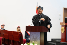 Mężczyzna, przewodniczący samorządu EDD, w czarnej todze i birecie przemawia przy mównicy, wykonując gest prawą ręką. W tle siedzą dwie kobiety i mężczyzna w akademickich strojach. Po prawej stronie widnieje napis „VERITATIS SPLENDOR”. 