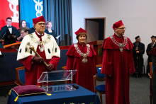 Trzy osoby w czerwonych togach i biretach stoi za przezroczystym pulpitem. Osoba po lewej - rektor, trzyma berło i ma togę z futrzanym kołnierzem. W tle widać osoby w czarnych strojach oraz czerwone i niebieskie flagi.