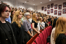 Młode osoby stojące w auli. Są to immatrykulowani studenci. Wszyscy są nieformalnie ubrani. Wszyscy patrzą w jednym kierunku. W tle na jasnej ścianie wiszą portrety w czerwonych ramkach
