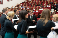 Chór w czasie występu. Sześć osób: trzy kobiety i trzech mężczyzn w eleganckich ubraniach. Kobiety noszą zielone spódnice, mężczyźni ciemne garnitury z zielonymi krawatami. Jedna kobieta trzyma dziecko. W tle publiczność siedzi na czerwonych krzesłach. 