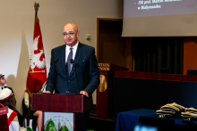 Doradca prezydenta. ubrany w ciemny garnitur, stoi za mównicą i przemawia. W tle widać flagę WUM.