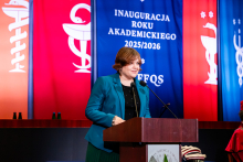 Kobieta, podsekretarz stanu w MNiSW w zielonej marynarce stoi przy mównicy i mówi do mikrofonu. W tle wiszą trzy banery: dwa czerwone i jeden niebieski z napisem „INAUGURACJA ROKU AKADEMICKIEGO 2025/2026” oraz skrótem „EFQS”. Banery zawierają symbole medyczne.