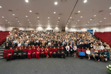 Uczestnicy immatrykulacji pozują do wspólnego zdjęcia. W pierwszym rzędzie siedzi 20 osób w czerwonych togach. Za nimi znajduje się kilkuset studentów w garniturach i sukienkach. Po prawej stronie sceny stoją osoby z szarfami. Wnętrze ma białe ściany i czerwone fotele. Uczestnicy patrzą przed siebie, mają poważne wyrazy twarzy. Zdjęcie pokazuje uroczystość akademicką.