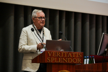 Mężczyzna w jasnej marynarce i okularach stoi za mównicą  wygłaszając przemówienie