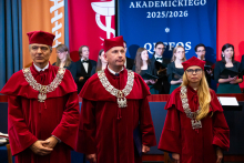 Trzy osoby w czerwonych togach i biretach stoją w sali. Są to prorektorzy. W tle widać czerwone i niebieskie flagi oraz osoby w czarnych strojach, które śpiewają lub grają. Na stole z niebieskim obrusem stoi przezroczysta skrzynka i leży poduszka.