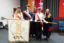 Pięć osób stoi w rzędzie na tle kolorowych flag. To poczet sztandarowy. Trzy osoby z przodu trzymają sztandar z napisem „Universitas Medica Varsoviensis 1809”. Osoby po bokach mają białe koszule, długie włosy i czapki z daszkiem. Osoba w środku nosi garnitur i krawat. Za nimi stoi dziekan.