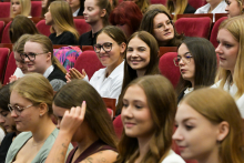 Grupa młodych osób w auli. Są to immatrykulowani studenci. Kilka z nich patrzy w obiektyw i się uśmiecha. Siedzą w czerwonych fotelach ustawionych w rzędach. 