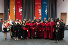 W auli pozuje grupa 20 osób w togach i biretach. Na środku stoi rektor w czerwonej todze z łańcuchem rektorskim. Po lewej stronie dwie osoby trzymają sztandar uczelni. W tle wiszą trzy banery: pomarańczowy, czerwony i niebieski, oznaczające wydziały WUM.