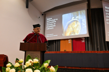 Mężczyzna w czerwono-czarnej todze i czarnym birecie, prof. Czajkowski, stoi przy mównicy i przemawia. W tle widzę dwa ekrany z prezentacją zatytułowaną „Wpływ technologii na zrozumienie choroby na przykładzie cukrzycy”. Na pierwszym planie leżą białe kwiaty.