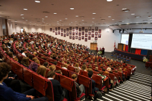 Aula wypełniona gośćmi immatrykulacji i studentami. Wszyscy siedzą na czerwonych fotelach i patrzą przed siebie, na środek auli, na której stoi prof. Czajkowski i daje wykład.