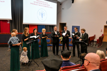 Chór w czasie występu. To 8 osób, ubranych w czarne garnitury i zielono-czarne suknie. Jedna z osób po lewej trzyma na rękach małe dziecko. Za nimi widać ekran multimedialny, przed nimi - słuchaczy.