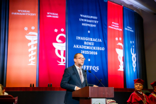 Mężczyzna w ciemnym garniturze, wicewojewoda mazowiecki, przemawia na środku auli. W tle wiszą kolorowe banery z nazwami wydziałów: Farmaceutycznego, Lekarskiego, Lekarsko-Stomatologicznego i Nauk o Zdrowiu. Po prawej stronie siedzą osoby w togach akademickich.