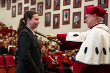 Rektor pasuje młodą kobietę na studentkę. W tle widać wypełnioną różnymi osobami aulę.