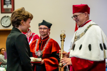 Młoda dziewczyna odbiera od rektora czapkę studencką. Obok rektora stoi dziekan Wydziału Farmaceutycznego w czarno-pomarańczowe todze.