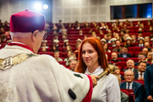 Na pierwszym planie młoda kobieta z rudymi włosami, ubrana formalnie. Jest w trakcie pasowania na studentkę. Rektor kładzie jej berło na ramieniu. W tle widać wypełnioną aulę.