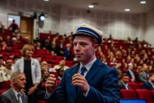 Młody mężczyzna, formalnie ubrany, stoi na środku sali i ma na głowie czapkę akademicką. W tle widać pełną aulę.