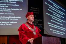 Mężczyzna w czerwonej todze i birecie, prorektor ds. nauki i transferu technologii, stoi za mównica i przemawia. W tle widać ekran multimedialny z wyświetlonymi słowami przyrzeczenia doktorskiego.