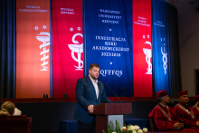 Młody mężczyzna, przewodniczący Samorządu Doktorantów, formalnie ubrany, stoi za mównicą i przemawia. W tle wiszą kolorowe banery z nazwami wydziałów i napisem „Inauguracja Roku Akademickiego 2025/2026”. 