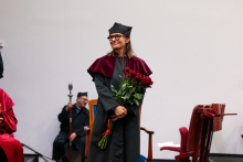 Kobieta w czarnej todze akademickiej stoi i trzyma bukiet czerwonych róż. Uśmiecha się i patrzy w prawo. W tle siedzi osoba w czerwonej todze, która trzyma berło. 