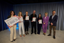 Siedem osób stoi w rzędzie na tle szarej zasłony. Pięć z nich trzyma w rękach certyfikaty. Kobieta po lewej stronie trzyma duży czek z napisem „Nagroda Publiczności” i kwotą 2000 zł.