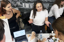 Cztery osoby stoją przy stole w jasnym pomieszczeniu. Na stole leży fantom niemowlęcia w jasnym ubraniu, obok butelka wody, ulotki i małe przedmioty medyczne. Na laptopie widać grafikę z rysunkiem dłoni i koszulki. Jedna osoba w białej koszulce gestykuluje, druga w czarnej bluzce stoi obok, a pozostałe osoby pochylają się nad stołem. 