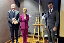 Trzy elegancko ubrane osoby: dwóch mężczyzn i kobieta pozują przy tablicy pamiątkowej prof. Kossakowskiego.