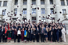 A large group of people on the steps in front of a monumental building with tall columns. The participants are dressed elegantly in suits and dresses. They all raise their hands and throw burgundy caps into the air.