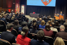 The auditorium during the graduation ceremony at Harvard Medical School. A person stands at the podium with the Harvard logo and addresses the participants. The participants sit in rows, and flags stand on the left side.