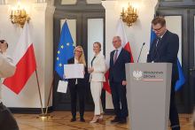 Trzy osoby stoją przed flagami Polski i Unii Europejskiej w eleganckiej sali, jedna trzyma otwarty dokument i torbę. Po prawej stronie znajduje się mównica z napisem „Ministerstwo Zdrowia”.