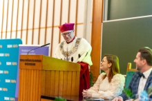 Osoba w ceremonialnym stroju akademickim, z czerwonym biretem i ozdobnym łańcuchem rektorskim, stoi przy drewnianej mównicy. Obok siedzą dwie osoby na zielonych krzesłach.