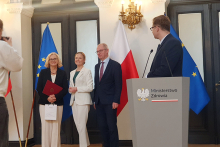 Trzy osoby stoją przed flagami Polski i Unii Europejskiej w eleganckiej sali, jedna trzyma bordową teczkę i torbę. Po prawej stronie znajduje się mównica z napisem „Ministerstwo Zdrowia”.