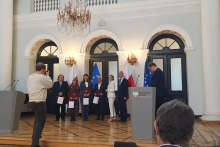 Sześć osób stoi w eleganckiej sali przed flagami Polski i Unii Europejskiej, trzy osoby trzymają bordowe teczki i białe torby. Po prawej stronie znajduje się mównica z napisem „Ministerstwo Zdrowia”, a po lewej fotograf robi zdjęcie.