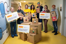 A group of people with large cardboard boxes and boards are standing in a bright hospital corridor. The boxes feature colorful graphics depicting woolen hats. 